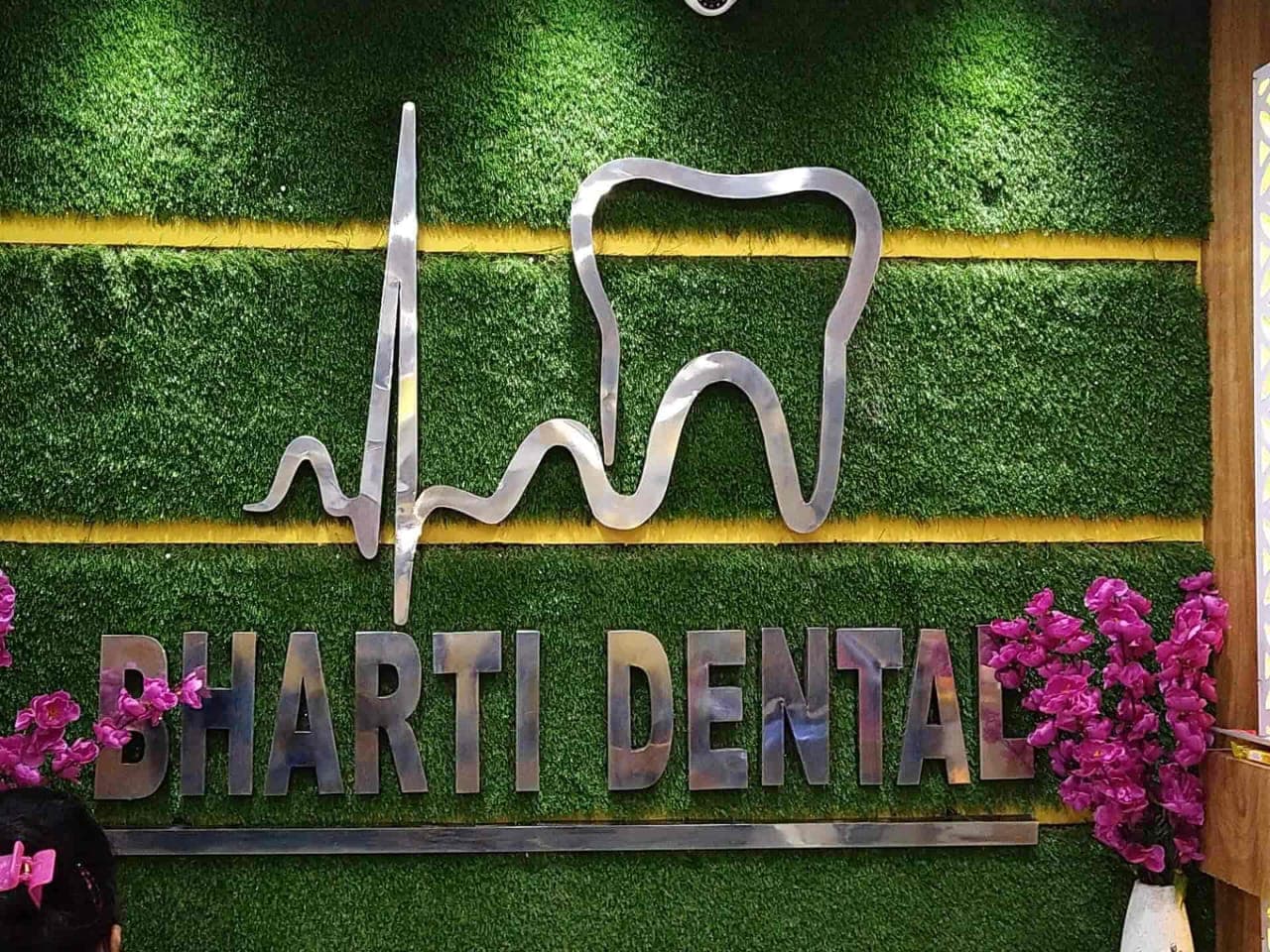 Silver Bharti Dental sign featuring a tooth heartbeat logo on a green grass wall.